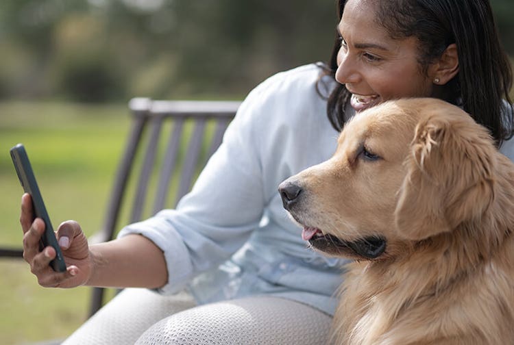 Smiling woman using her phone with her pet golden retriever sitting on a bench outdoors